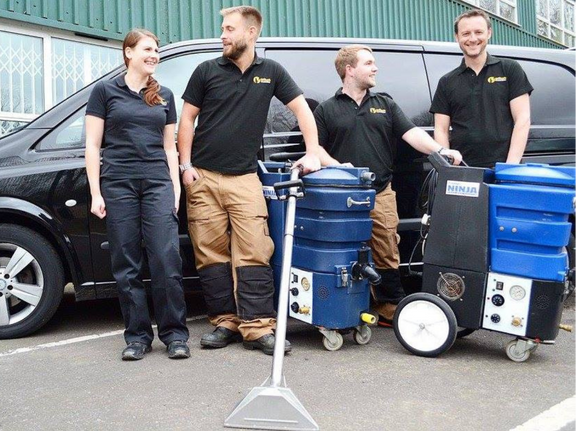 This is a photo of Mill Hill Carpet Cleaning carpet cleaners (three men and one woman) standing in fromt of their black van, with two steam cleaning carpet machines next to them